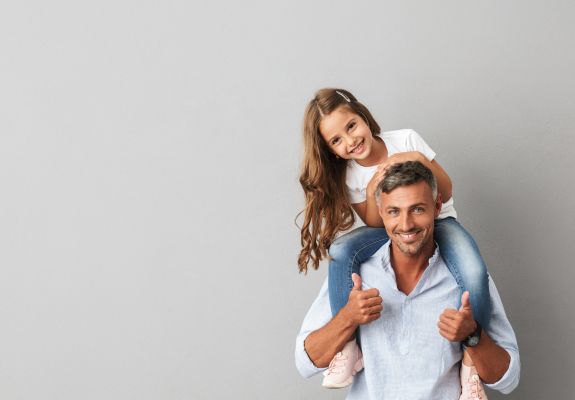 Tochter sitzt lachend beim Vater auf den Schultern, der Daumen hoch gibt