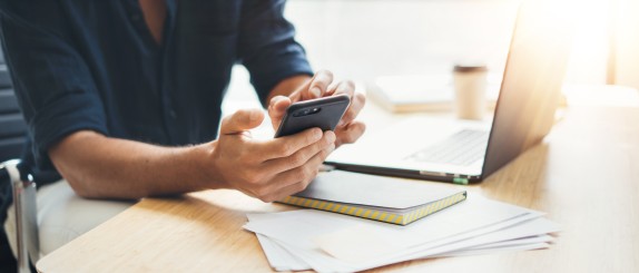 Mann sitzt beim Arbeiten am Schreibtisch und schaut etwas auf seinem Handy nach