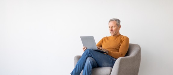 Ein älterer Herr sitzt in einem Sessel und ließt am Laptop