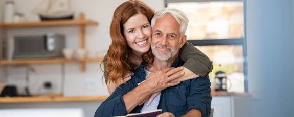 Älterer Mann sitzt am Esstisch und seine Frau umarmt ihn liebevoll von hinten