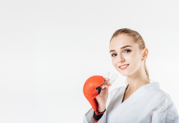 Junge Frau im Kampanzug mit Boxhandschuhen zeigt lächelnd ihren Mundschutz